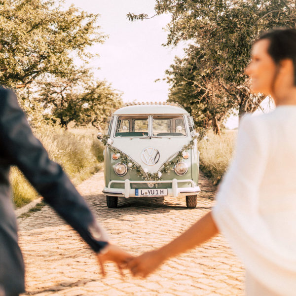 Hochzeitspaar Hand in Hand vor VW Bulli Oldtimer