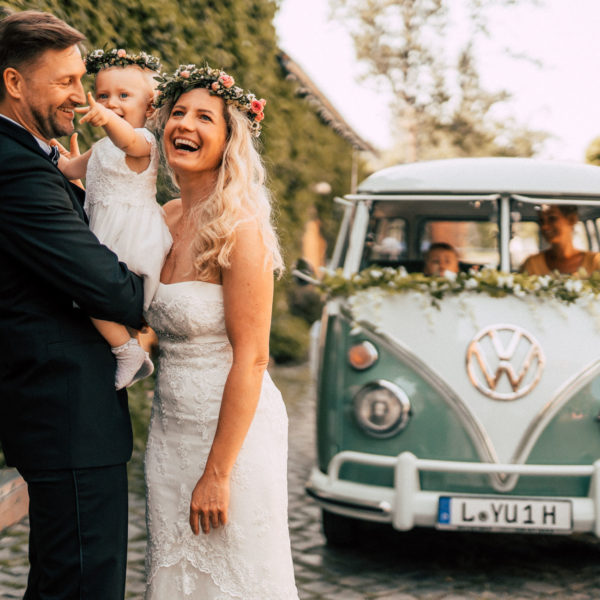 Chauffeurin Nancy Tschörner in ihrem VW Bulli Oldtimer im Hintergrund, vorne Hochzeitspaar mit Kind